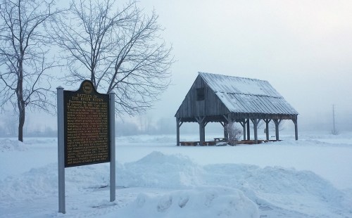 A snowy landscape featuring a historical marker and a rustic wooden shelter under a foggy sky.