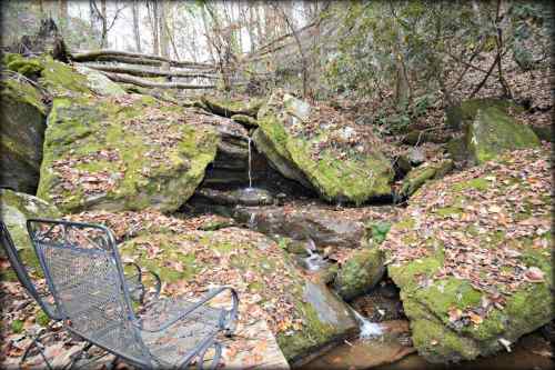 A serene scene of moss-covered rocks with a small stream, surrounded by autumn leaves and a metal chair nearby.