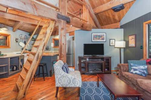 Cozy cabin interior with wooden beams, a staircase, and a living area featuring a TV, sofa, and armchair.