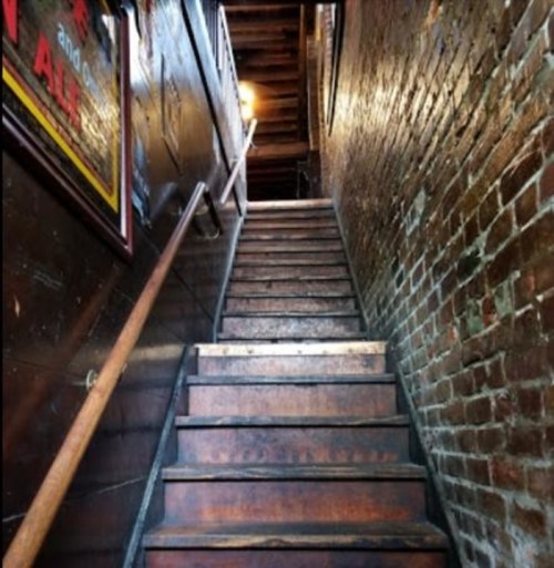 A dimly lit wooden staircase leading upward, flanked by brick walls and a handrail.