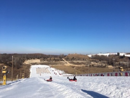 A snowy hill with people tubing down, surrounded by a clear blue sky and distant hills.