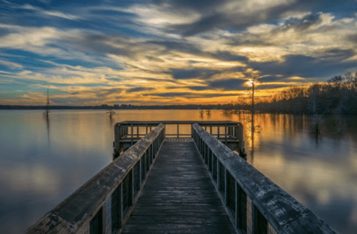 A serene wooden pier extends into a calm lake at sunset, with colorful clouds reflecting on the water's surface.
