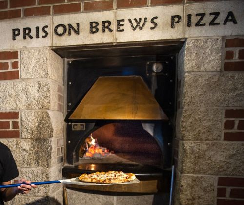 A person holds a pizza paddle in front of a brick oven labeled "Prison Brews Pizza," with flames visible inside.