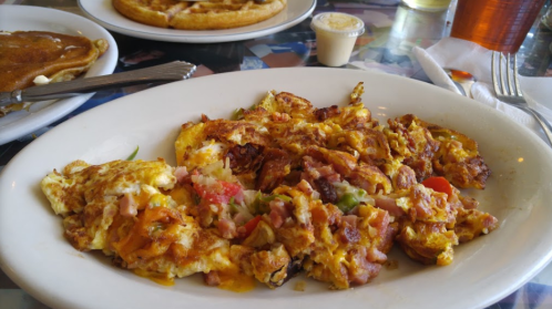 A plate of scrambled eggs mixed with diced vegetables and ham, served with a side of syrup and a waffle in the background.