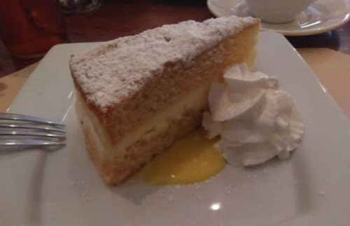 A slice of cake dusted with powdered sugar, served with whipped cream and a drizzle of lemon sauce on a white plate.