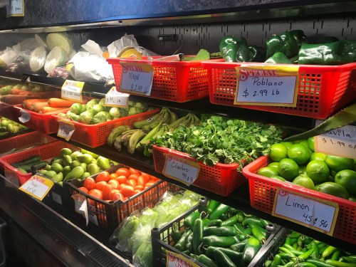 A colorful display of fresh vegetables and herbs in baskets, with price tags, in a grocery store.