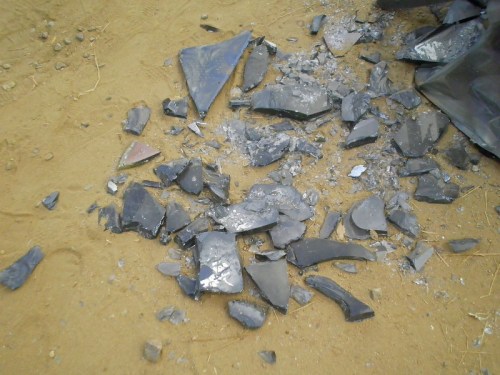 Scattered shards of dark, broken glass on a sandy surface.