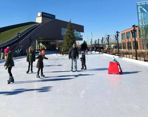 A group of people ice skating on a sunny day, with a building and a hill in the background.