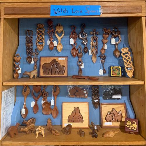 A display of intricately carved wooden love spoons and animal figures on shelves, labeled "Welsh Love Spoons."