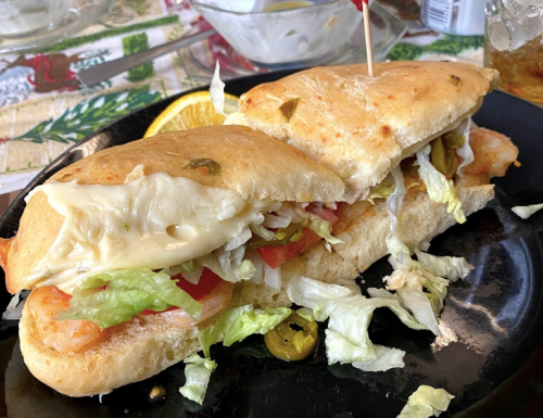 A close-up of a sandwich filled with shrimp, lettuce, tomato, and cheese, served on a black plate with a slice of orange.
