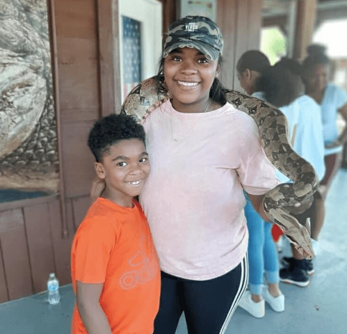 A woman smiles while holding a snake around her neck, standing next to a boy in an orange shirt.