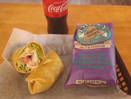 A wrap filled with lettuce, tomato, and other ingredients, alongside a bag of salt and vinegar potato chips and a Coke.