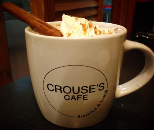 A large mug from Crouse's Cafe filled with whipped cream, a cinnamon stick, and a sprinkle of crumbs on top.