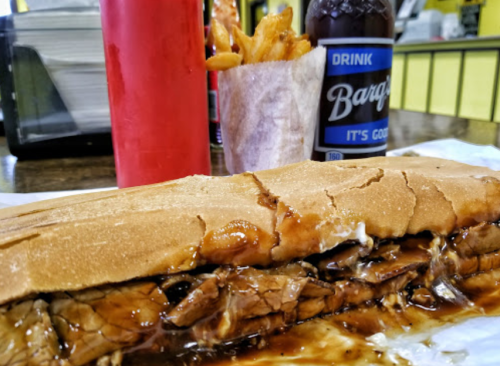 A close-up of a sandwich filled with meat and sauce, accompanied by fries and a bottle of Barq's soda in a diner setting.
