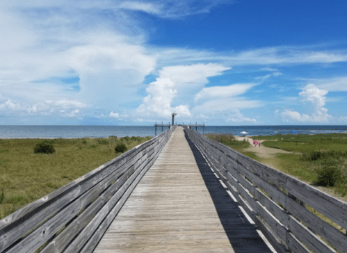 A wooden pier extends into the ocean under a bright blue sky with fluffy clouds and green grass on either side.