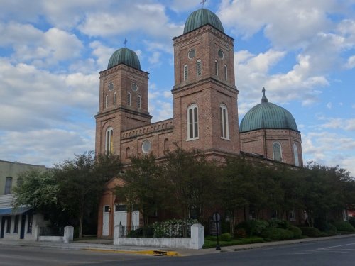 Historic brick church with two towers and domed roofs, surrounded by trees and a cloudy blue sky.