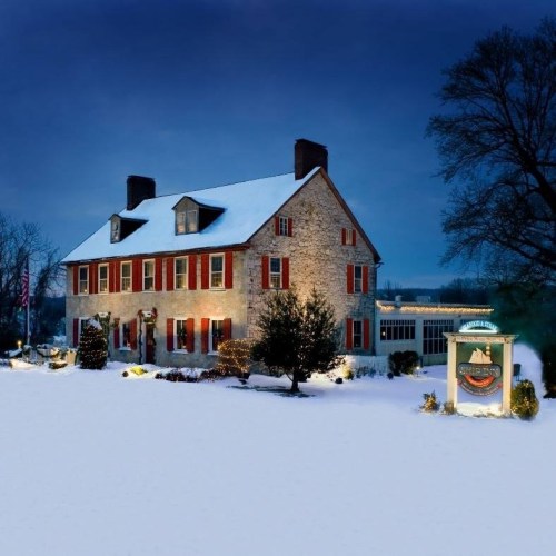 A charming stone house with red shutters, surrounded by snow, illuminated by festive lights in a winter evening setting.