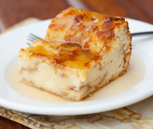 A slice of bread pudding drizzled with syrup on a white plate, resting on a patterned cloth.