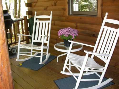 Two white rocking chairs and a small table with a potted flower on a wooden porch surrounded by trees.