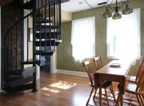 A dining area with a wooden table and chairs, featuring a spiral staircase and large windows with sheer curtains.