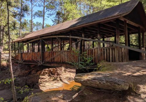 A wooden covered bridge spans a rocky creek, surrounded by trees and sunlight filtering through the leaves.