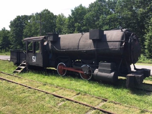 A vintage steam locomotive parked on grass, featuring a black body and red wheels, surrounded by trees.