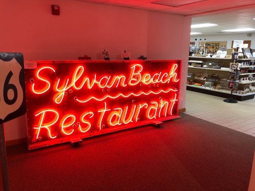Neon sign for Sylvan Beach Restaurant in a hallway, with a display of items visible in the background.