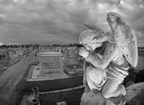 A stone angel statue kneels in prayer, overlooking a cemetery filled with gravestones under a cloudy sky.