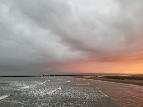 A dramatic seascape at sunset, with dark clouds and vibrant orange hues reflecting on the water. Waves gently roll in.
