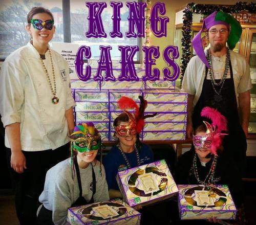 A group of four people in festive masks and beads pose with king cakes, celebrating Mardi Gras.