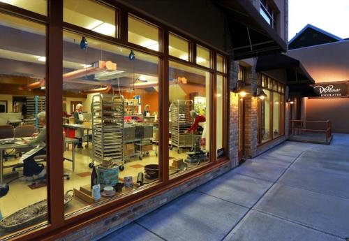 A chocolate shop with large windows showing workers inside, surrounded by baking trays and equipment, at dusk.