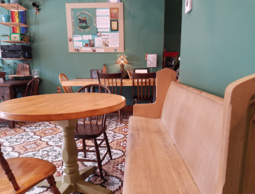 A cozy café interior featuring a wooden table, chairs, and a bench against a green wall with decorative elements.