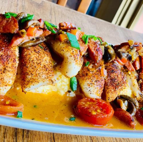 A plate of seasoned fish fillets topped with sautéed vegetables and herbs, served over a bed of creamy sauce.