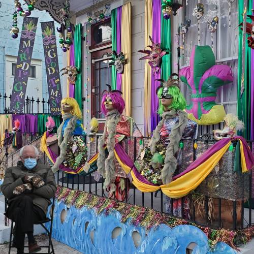 A colorful Mardi Gras display with masked figures and vibrant decorations, while a person sits nearby wearing a mask.