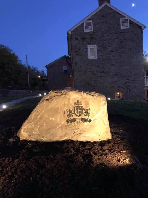 A large illuminated rock with a crest design, set in a landscaped area near a stone building at dusk.