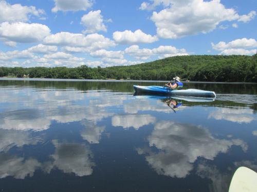 A person kayaking on a calm lake, surrounded by lush greenery and fluffy clouds reflected in the water.