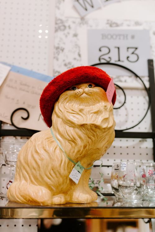 A ceramic cat figurine wearing a red hat, displayed on a shelf with various items in a vintage shop.