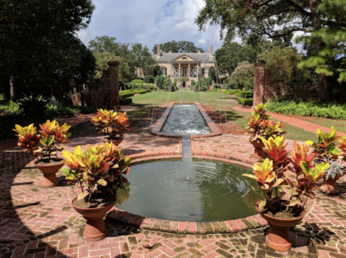A landscaped garden with a central pond, colorful plants, and a fountain, leading to a grand mansion in the background.