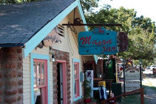 A quaint storefront with a colorful sign reading "The Mason Jar," surrounded by trees and outdoor decor.