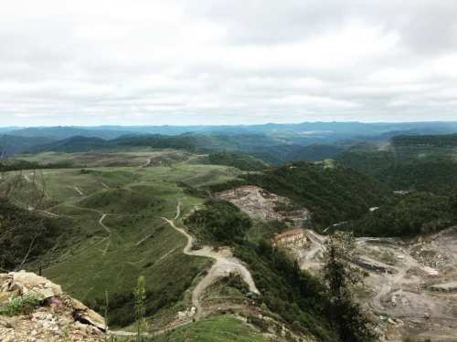 A panoramic view of rolling green hills and valleys, with visible mining activity in the foreground under a cloudy sky.