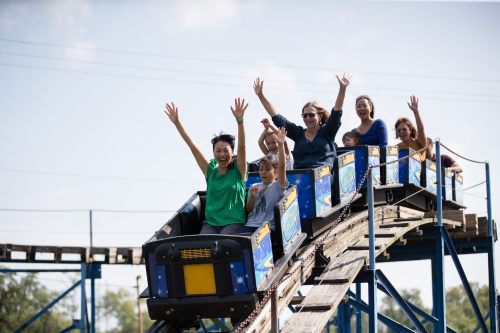 A group of people joyfully riding a roller coaster, raising their hands in excitement against a clear blue sky.