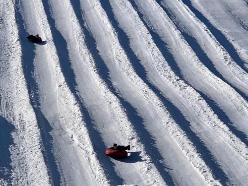 Two people sledding down a snowy slope, leaving behind wavy tracks in the fresh snow.