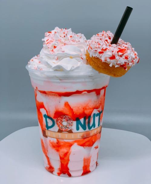 A festive milkshake topped with whipped cream, red syrup, and a donut, served in a clear cup with a straw.
