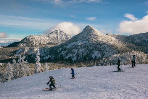 Skiers navigate a snowy slope with mountains and trees in the background under a clear blue sky.