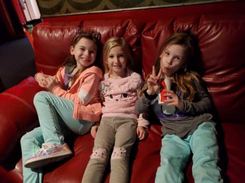 Three young girls sit on a red couch, smiling and posing playfully, dressed in colorful outfits.