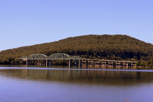 A green bridge spans a calm lake, with a wooded hill rising in the background under a clear blue sky.