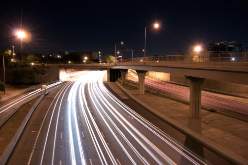 Long exposure shot of a highway at night, showcasing light trails from moving vehicles under a bridge.