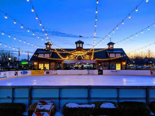 A winter ice skating rink at dusk, adorned with string lights and a charming building in the background.