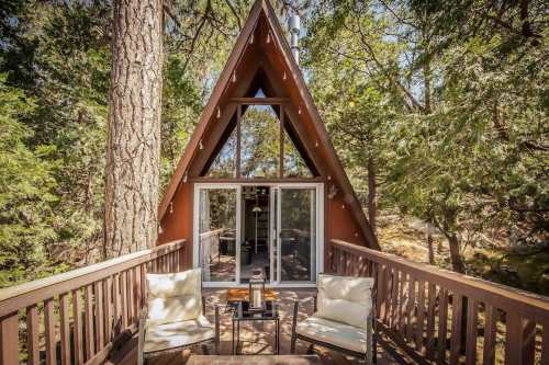 A cozy A-frame cabin surrounded by trees, featuring a deck with two chairs and a small table.