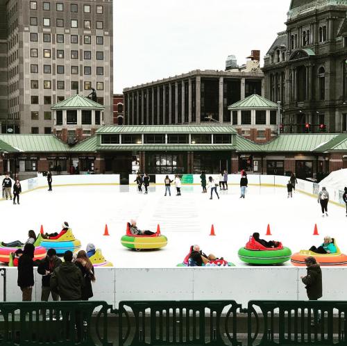 A winter scene with people ice skating and others sitting in colorful inflatable tubes on an outdoor rink.